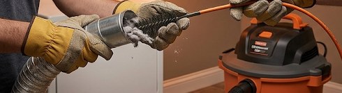 Hands in work gloves using flexible rod brush to clean a dryer vent duct, with lint debris being removed